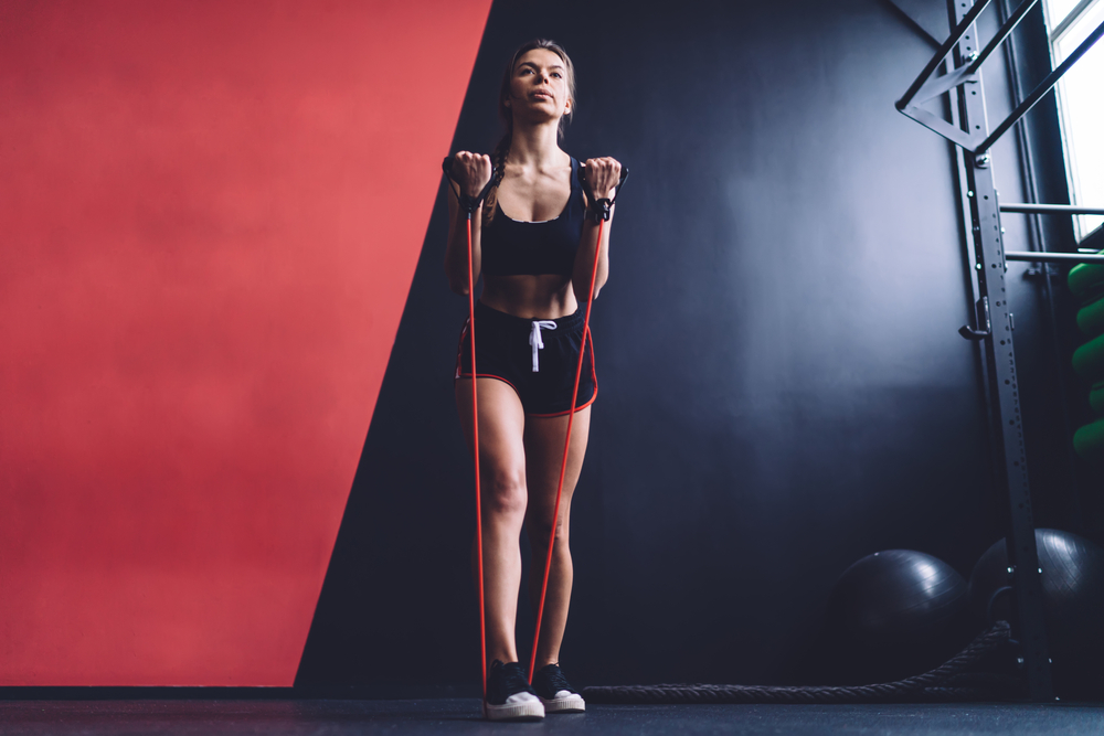 Female concentrated athlete in sportswear with perfect sportive figure working out with band upright rows standing in fitness gym looking away