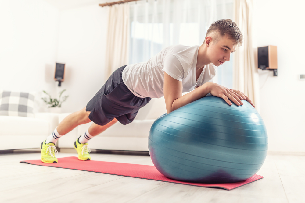 Working out at home by a young man holding a plank position using a blue fit ball and red mat inside a living room.