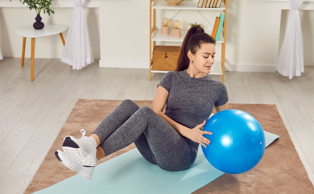 Young slim woman doing fit exercises lying on yoga mat in the living room at home using fit ball. Sporty girl exercising indoors. Healthy lifestyle, fitness and workout sport concept. This exercise targets your obliques, helping to sculpt your waistline.