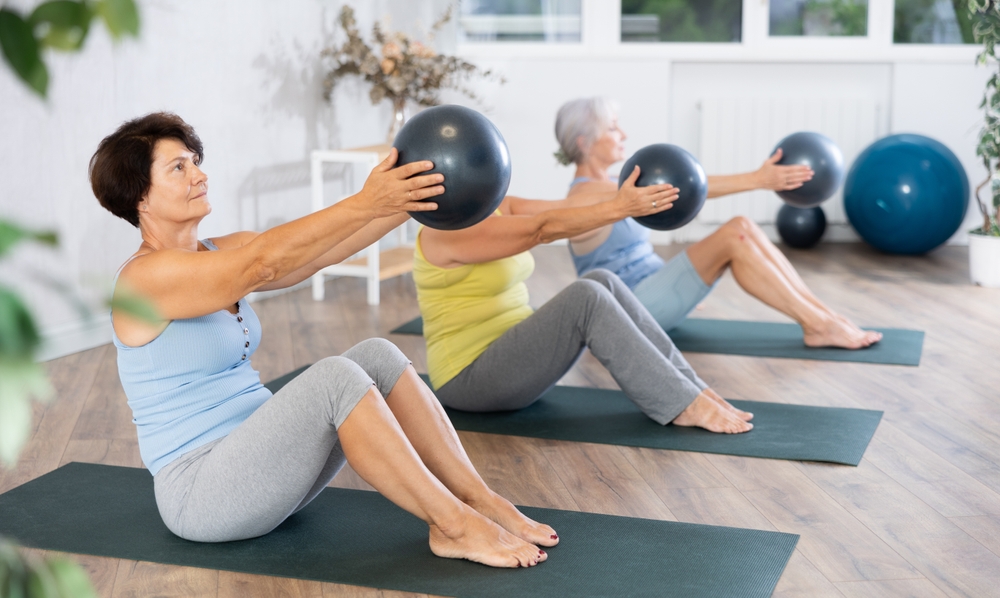 Calm mature woman doing pilatess sitting on black mat with soft ball in hands during workout session